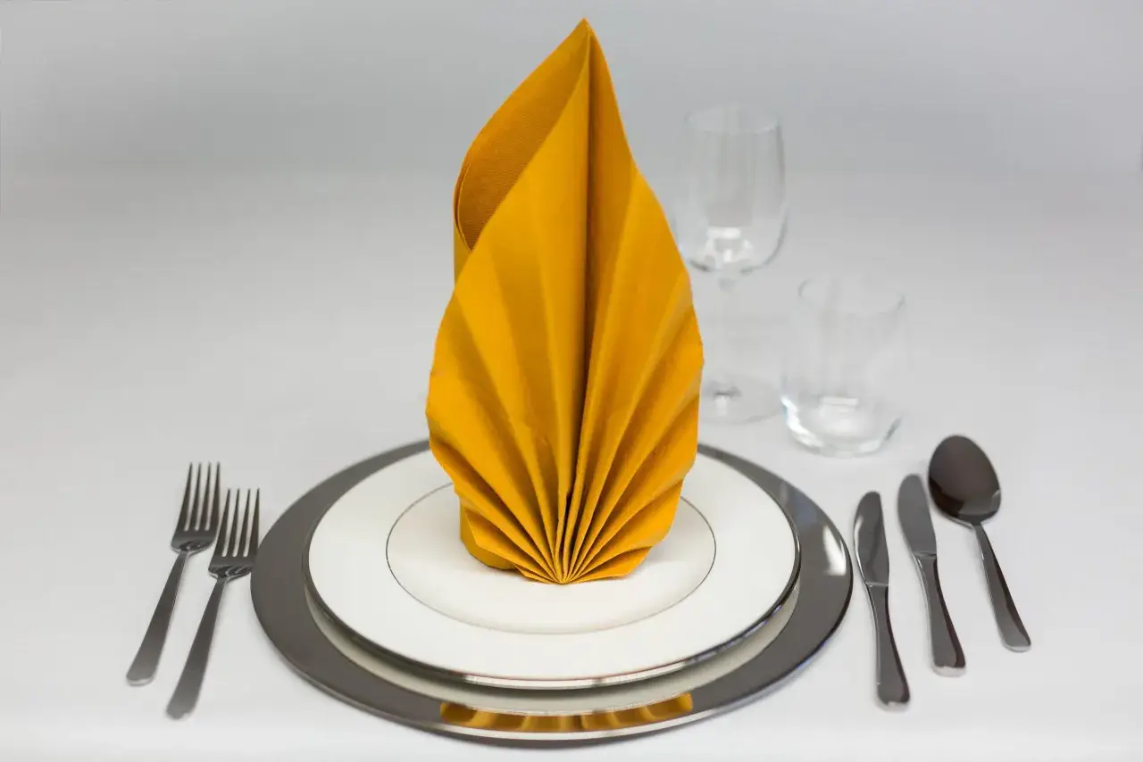 Ein festlich gedeckter Tisch mit einem gelben, gefalteten Servietten-Fächer. Gedeck mit Tellern, Besteck und Gläsern.