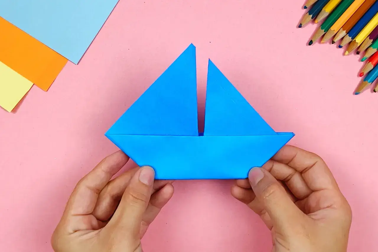Hände falten ein blaues Papierboot. Bunte Stifte und Papier liegen bereit für weitere Bastelprojekte.