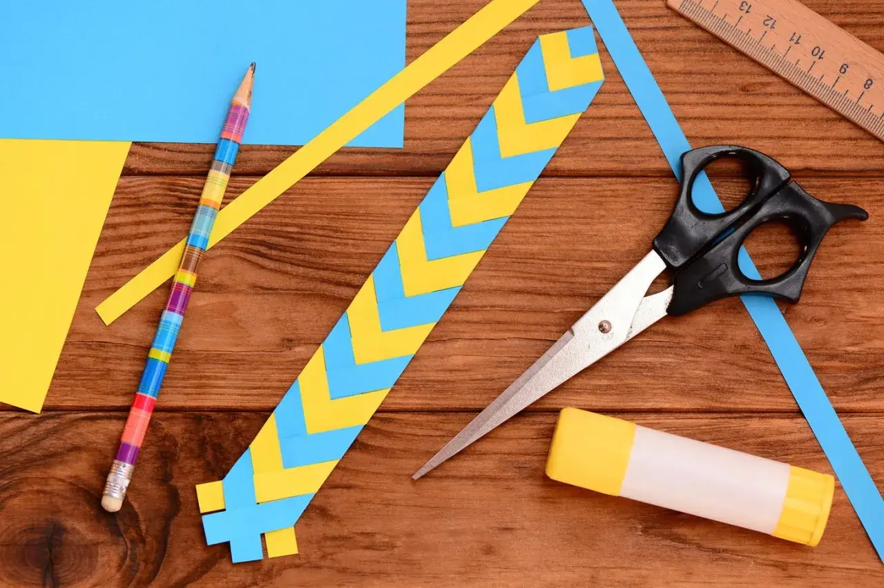 Bunte Streifen aus Papier werden zu einem Lesezeichen gebastelt. Schere, Klebestift und ein farbiger Bleistift liegen bereit.