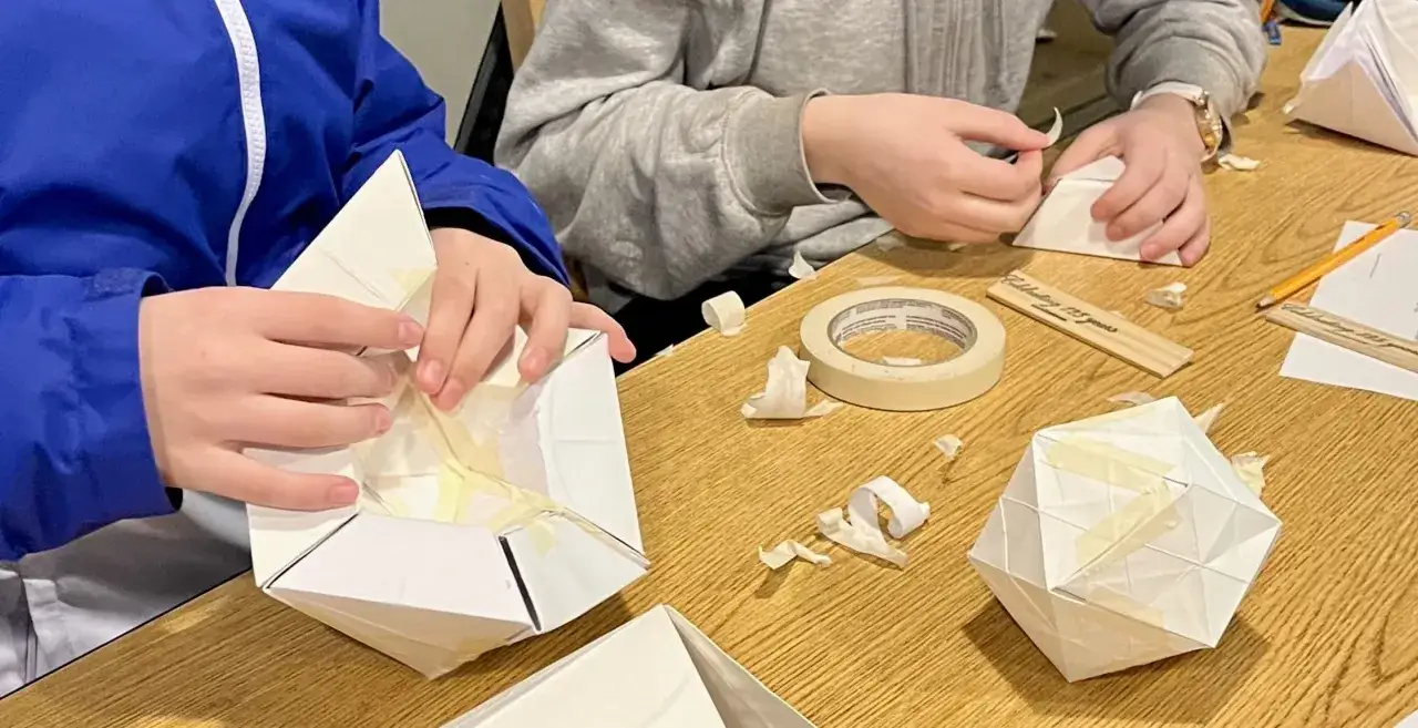 Kinder falten bei einem Origami-Kurs Papierfiguren. Klebeband und Papierfetzen liegen auf dem Tisch.