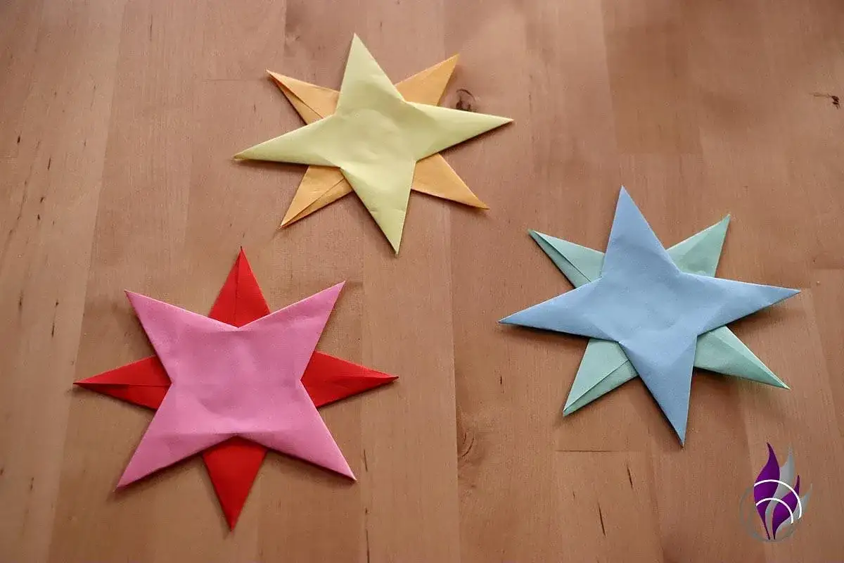 Drei bunte Papiersterne liegen auf einem Holztisch. Sie sind das Ergebnis von Wurfstern basteln.