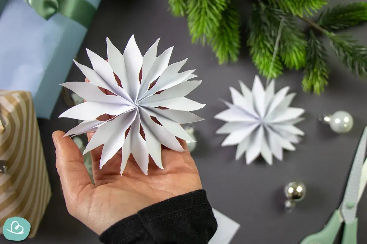 Hand h&auml;lt selbstgemachten DIY Christbaumschmuck aus Papier. Daneben liegen Geschenke, Tannenzweige und Kugeln.