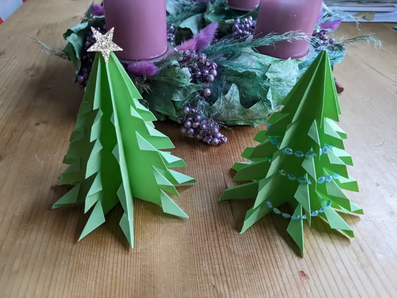 Zwei gr&uuml;ne origami tannenbaum flach stehen vor einem Adventskranz mit lila Kerzen. Einer ist mit einer goldenen Sternspitze verziert.