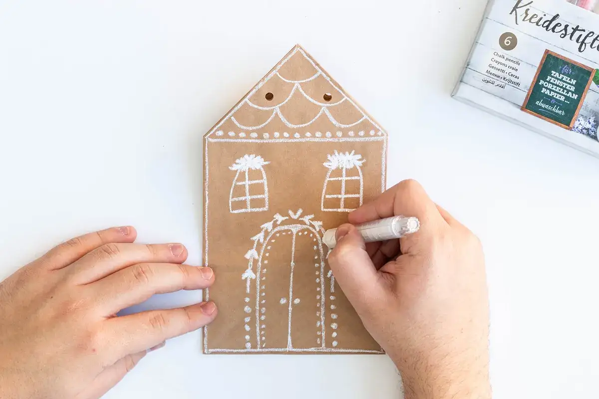 Mit wei&szlig;en Kreidestiften werden weihnachts giveaways selbstgemacht. Ein Haus aus braunem Papier wird verziert.