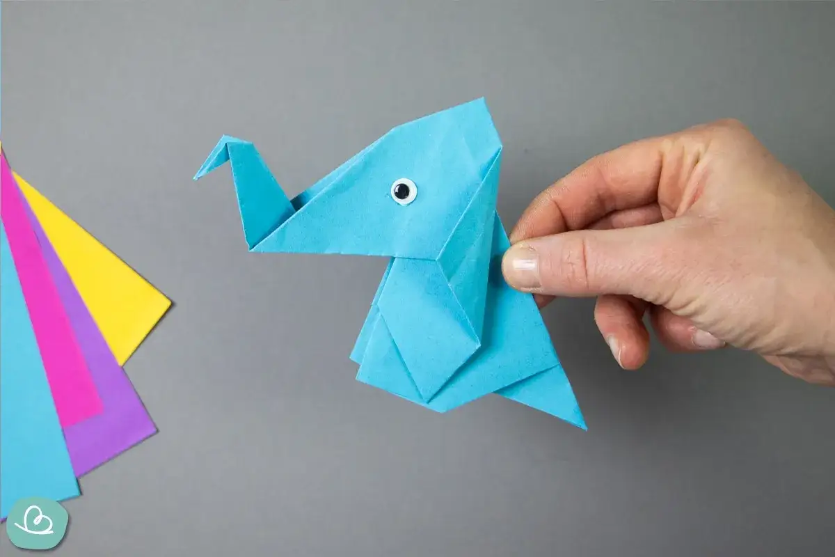 Ein blauer origami elefant mit einem kleinen Knopfauge wird von einer Hand gehalten. Daneben liegen bunte Papierb&ouml;gen.