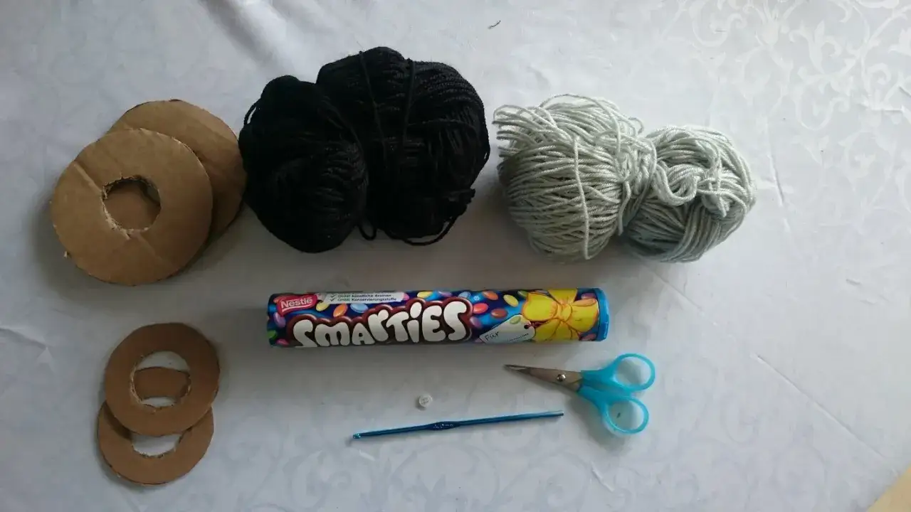 Materialien f&uuml;r ein Geldgeschenk: Pappringe, schwarzes und hellgraues Garn, Smarties-Rolle, Knopf, H&auml;kelnadel und Schere. So l&auml;sst sich eine Hantel basteln.