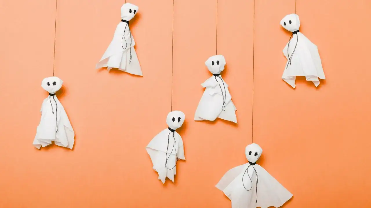 Selbstgemachte Halloween Deko: Papiergeister schweben vor orangefarbenem Hintergrund. Einfache Bastelideen mit Vorlagen.