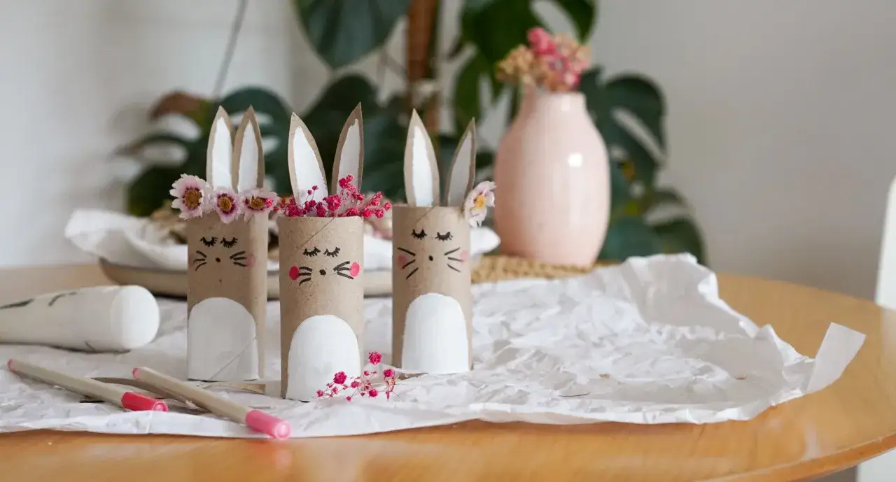 Drei süße Hasen aus Klopapierrollen, dekoriert mit Blumen und Stiften daneben.