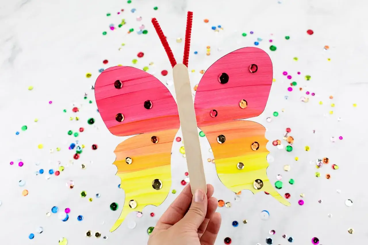 Bunte, glitzernde Schmetterlinge zum Basteln, gehalten in einer Hand, umgeben von Konfetti.
