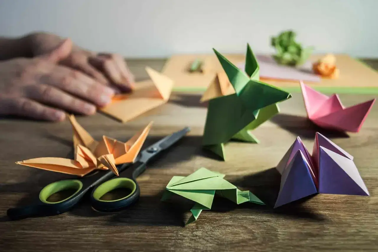 Bunte Papierfiguren, wie Kr&auml;ne und Fr&ouml;sche, liegen neben einer Schere. Eine Hand faltet Papier, als w&uuml;rde sie eine neue origami vorlage erstellen.