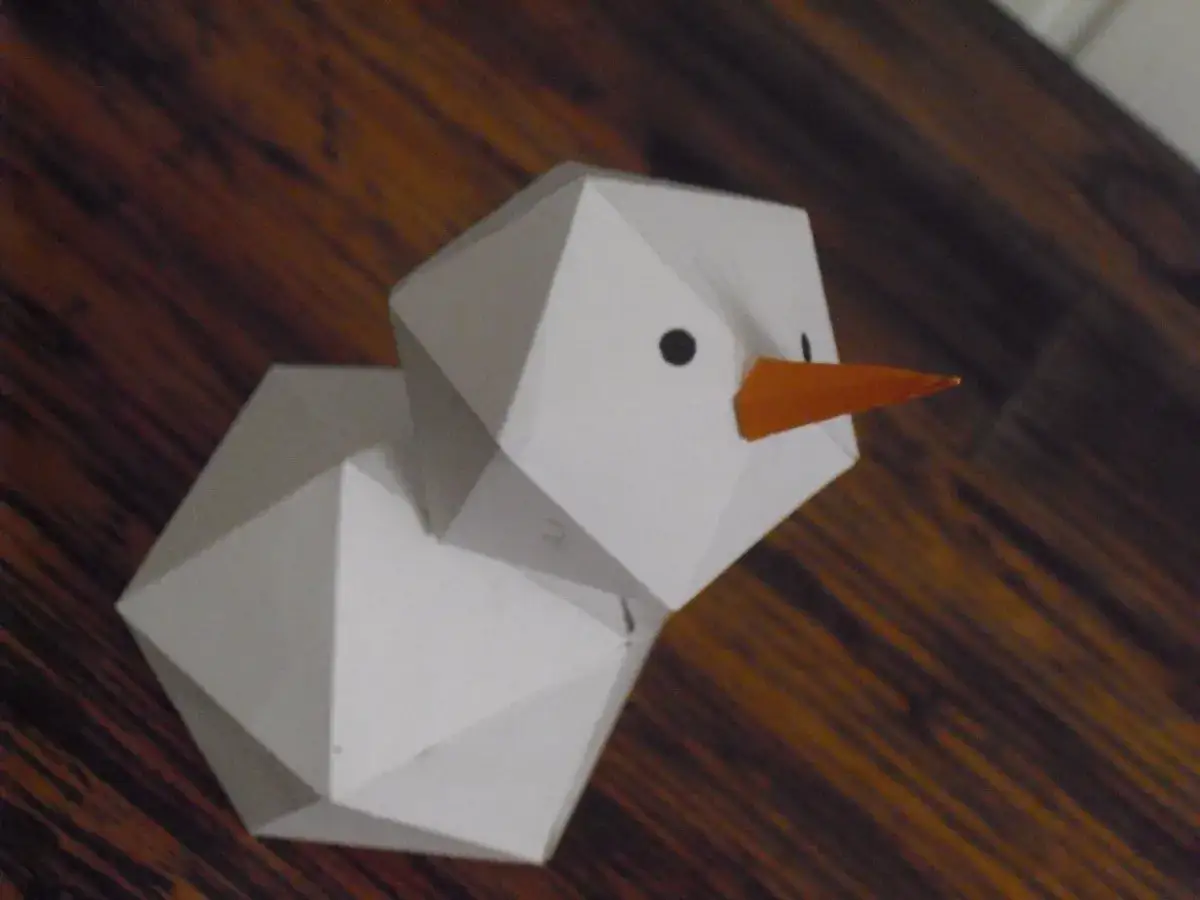 Geometrischer Schneemann, gefaltet aus Papier, mit orangefarbener Karotte als Nase und schwarzen Augen.