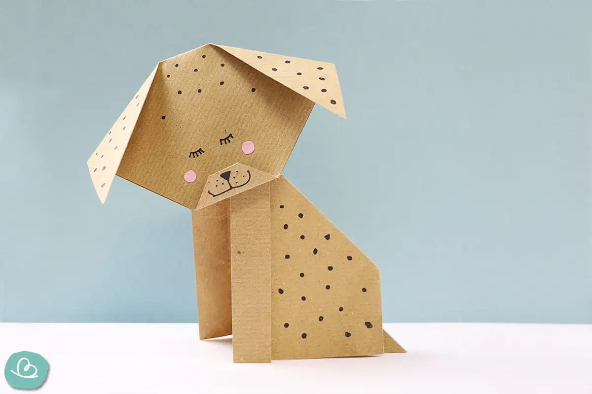 Ein süßer falten hund aus braunem Papier mit schwarzen Punkten und rosa Wangen sitzt auf einem weißen Tisch vor hellblauem Hintergrund.