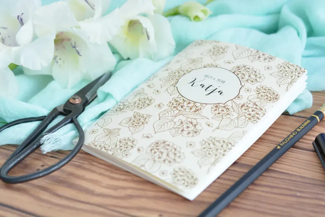 Ein Notizbuch mit Blumenmuster, Schere und Stift &ndash; bereit f&uuml;r kreatives buch basteln. Wei&szlig;e Blumen und mintfarbener Stoff im Hintergrund.