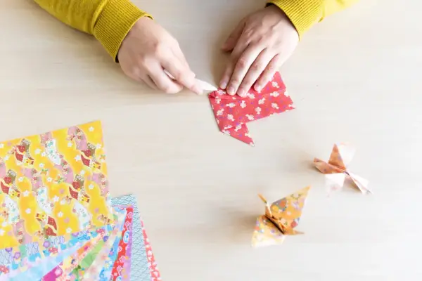 Hände falten Papier für eine Origami Kugel Anleitung. Mehrere bunte Origami-Papierbögen liegen bereit.