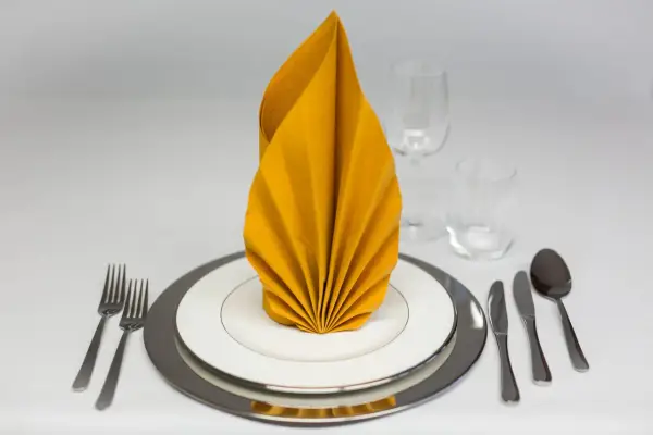 Ein festlich gedeckter Tisch mit einem gelben, gefalteten Servietten-Fächer. Gedeck mit Tellern, Besteck und Gläsern.