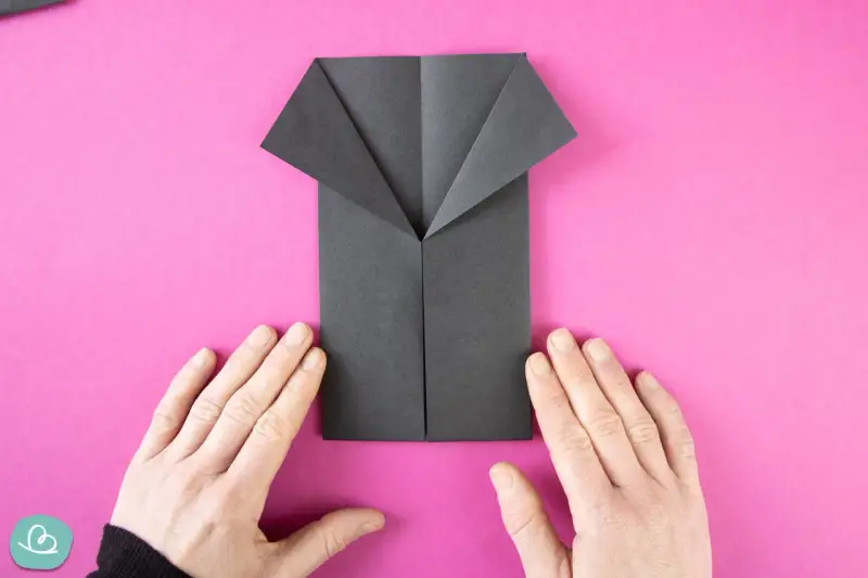 Ein schwarzes Origami-Hemd wird auf pinkem Hintergrund gefaltet. Hände halten das Papier.
