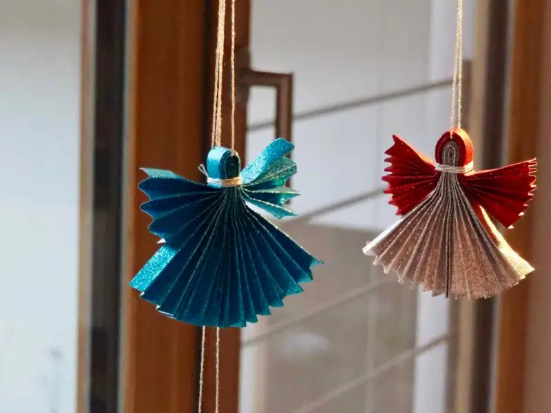 Zwei glitzernde Origami Weihnachts Engel, einer in Blau, der andere in Rot und Silber, hängen an Fäden.